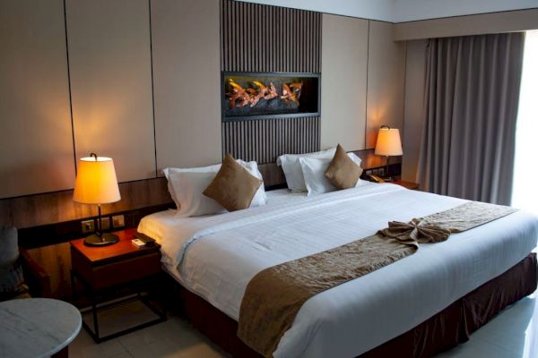 A cozy hotel room with a large bed, decorative pillows, lamps, a wall-mounted display, and curtains, creates an inviting atmosphere.