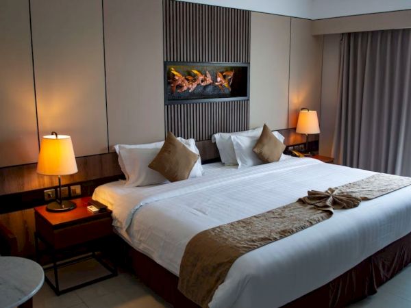 A cozy hotel room with a large bed, decorative pillows, lamps, a wall-mounted display, and curtains, creates an inviting atmosphere.