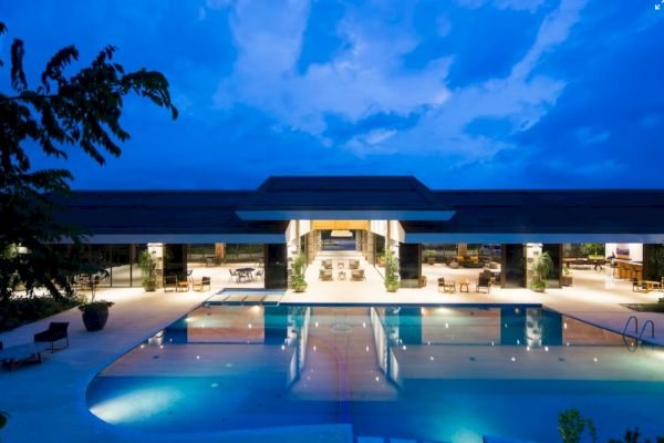 The image shows a modern building with a large swimming pool in front, under a twilight sky with scattered clouds.
