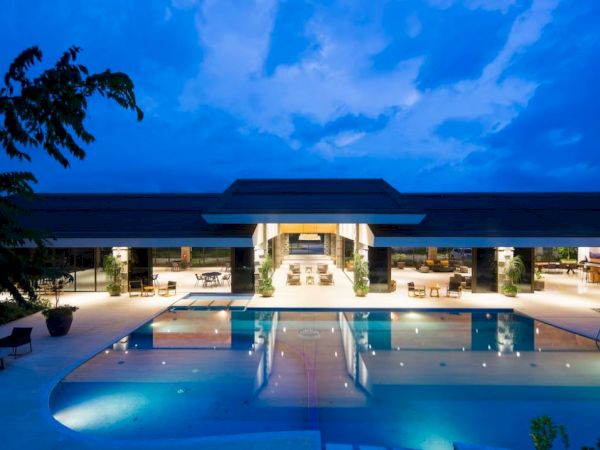 The image shows a modern building with a large swimming pool in front, under a twilight sky with scattered clouds.