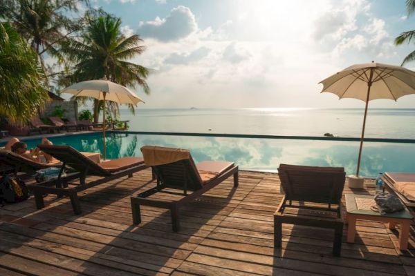 Wooden deck with lounge chairs and umbrellas overlooking a pool and ocean. Tropical palm trees frame the serene, sunlit scene.