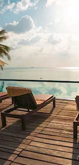 Wooden deck with lounge chairs and umbrellas by an infinity pool, overlooking the ocean with palm trees and a bright sky.