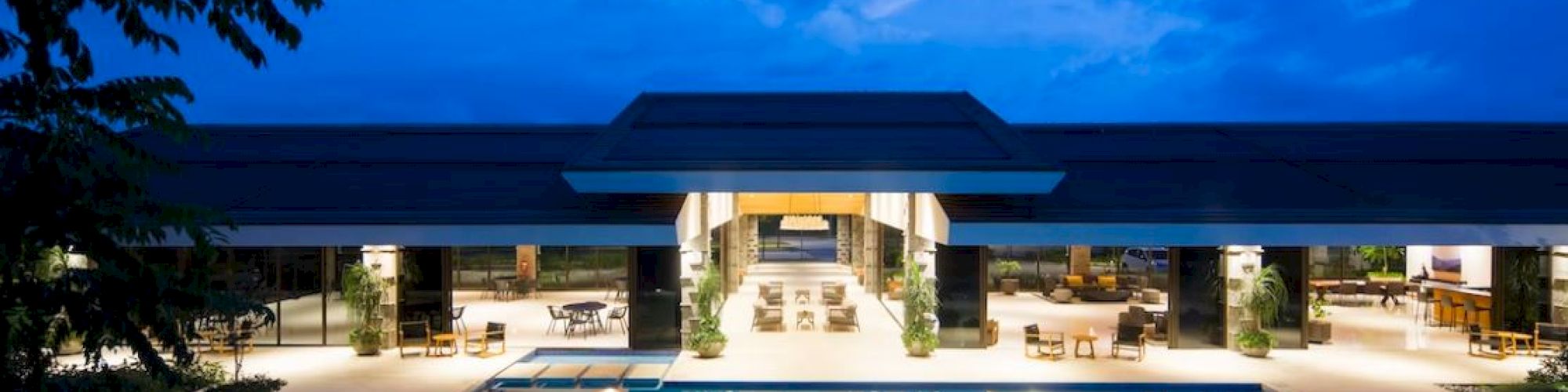 A luxurious building at night with a large illuminated pool in the foreground and seating areas inside.