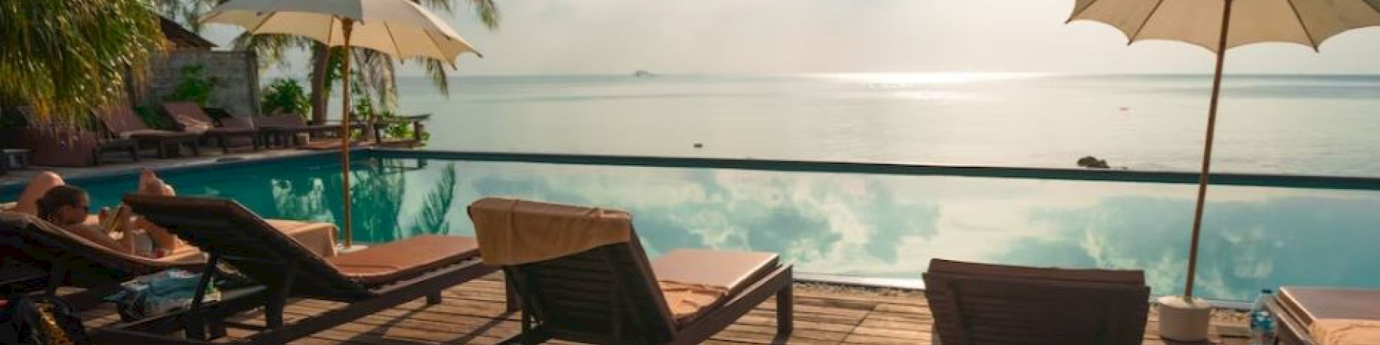 A serene beachfront scene with lounge chairs, umbrellas, and a calm horizon over a pool, surrounded by palm trees under a bright sky.