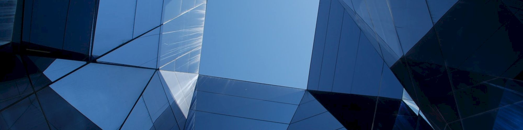 The image shows a geometric architectural structure with reflective glass panels and a clear blue sky visible through the center opening.