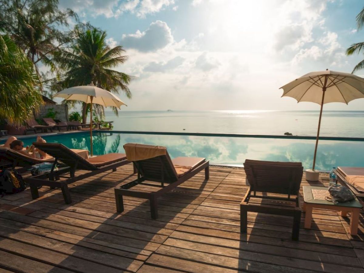 Poolside scene with lounge chairs, umbrellas, and palm trees by the water under a bright, cloudy sky.