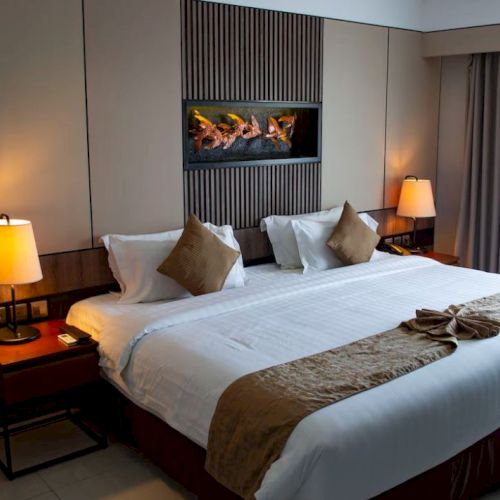 A well-lit hotel room featuring a large bed with brown and beige decor, bedside tables with lamps, and a wall art piece above the bed.