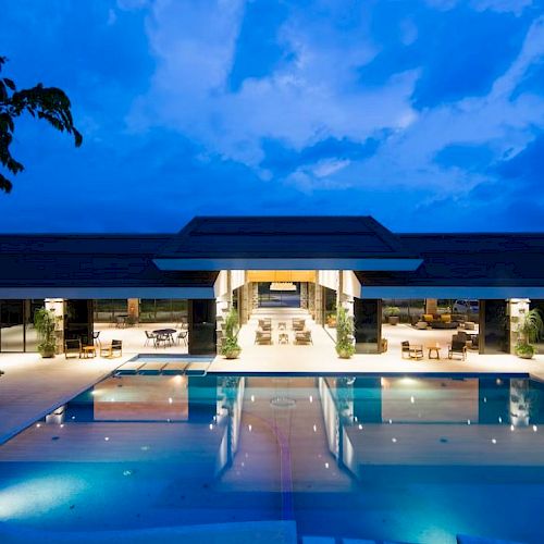 A modern building with large glass doors and outdoor seating overlooks a well-lit swimming pool at dusk, surrounded by trees.