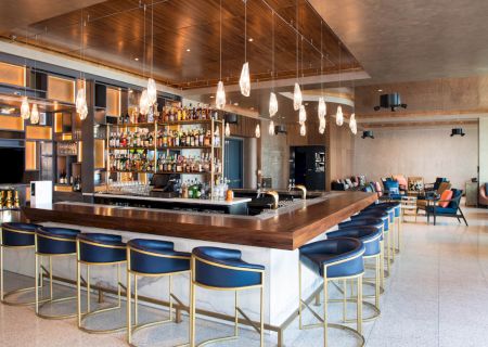 A modern bar interior with a sleek wooden counter, blue chairs, hanging lights, and a well-stocked bar.
