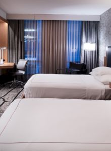 The image shows a modern hotel room with two beds, a desk, chair, TV, and stylish decor.