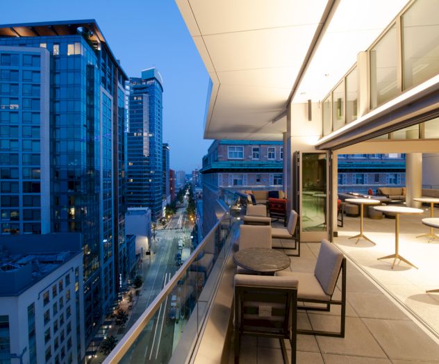 A modern city balcony at dusk with outdoor seating. Tall buildings line the illuminated street below, creating an urban atmosphere.