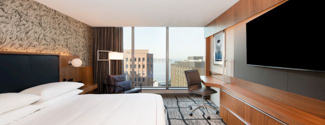 A modern hotel room features a large bed, a flat-screen TV, a work desk, and a window with a city view and natural light.