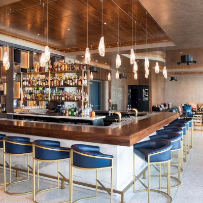 A modern bar with a sleek design, featuring a well-stocked bar, blue stools, and stylish pendant lighting in a spacious lounge area.