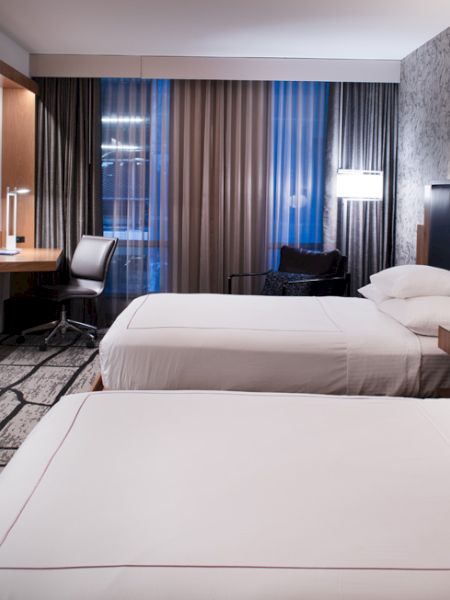 A modern hotel room with two beds, a wall-mounted TV, desk, chair, and window with curtains, decorated in neutral tones and patterns.