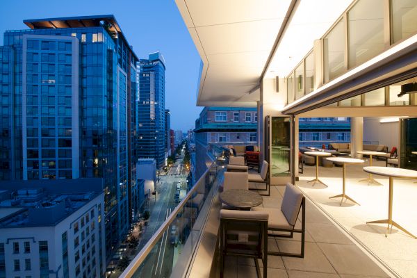 A modern cityscape at dusk with a high-rise building view from a stylish balcony featuring seating and tables, overlooking the lit street below.