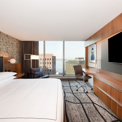A modern hotel room with a large bed, wall-mounted TV, desk, chair, and a window showing a cityscape view and body of water.