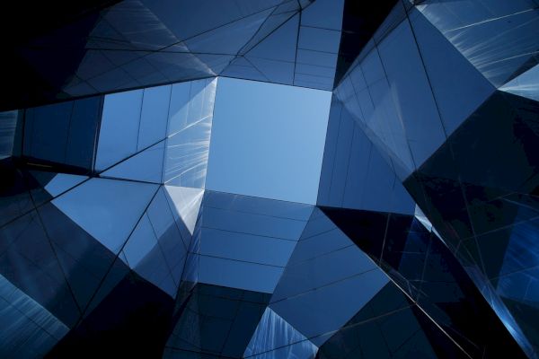 A view of the sky through geometric, reflective glass structures, creating an intricate, angular pattern with blue and dark hues.