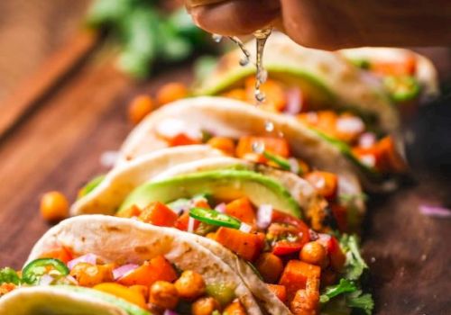 Tacos filled with chickpeas, vegetables, and garnished with herbs and avocado slices, with lime being squeezed over them.
