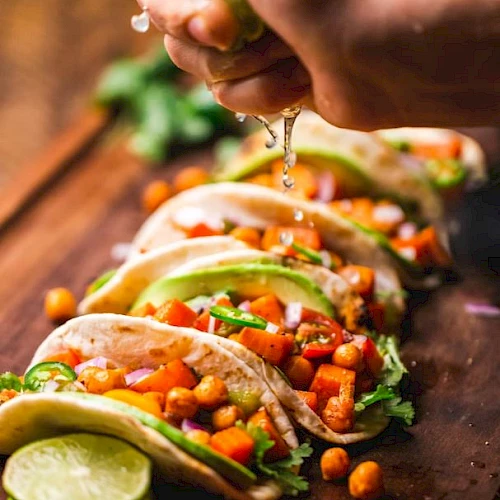 Tacos filled with chickpeas, vegetables, and garnished with herbs and avocado slices, with lime being squeezed over them.