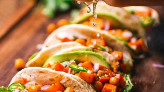 Tacos filled with chickpeas, vegetables, and garnished with herbs and avocado slices, with lime being squeezed over them.