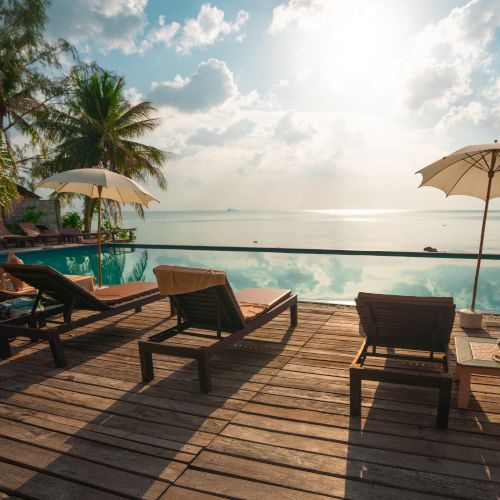 A serene tropical scene with lounge chairs, umbrellas, a pool, and a view of the ocean under a bright sky surrounded by palm trees.