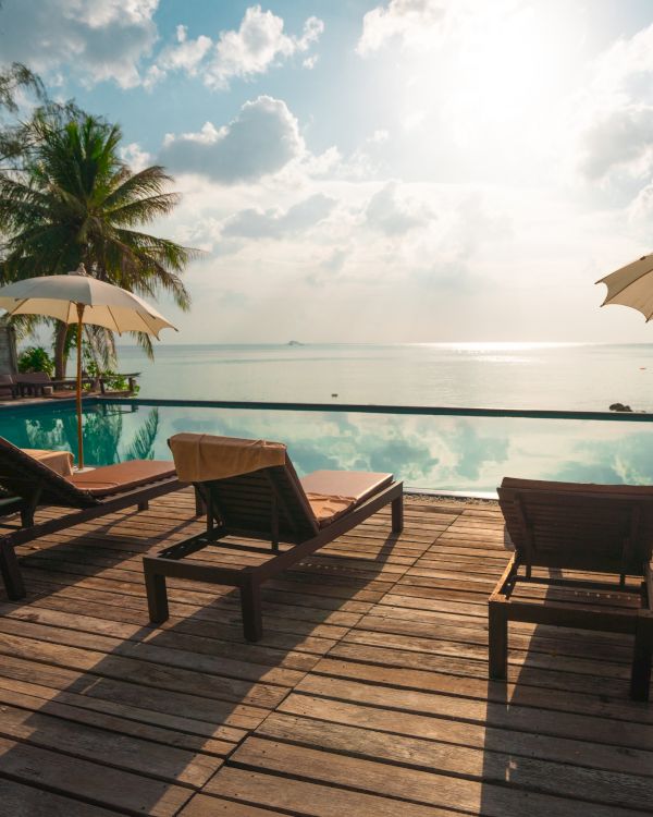 A serene tropical scene with lounge chairs, umbrellas, a pool, and a view of the ocean under a bright sky surrounded by palm trees.