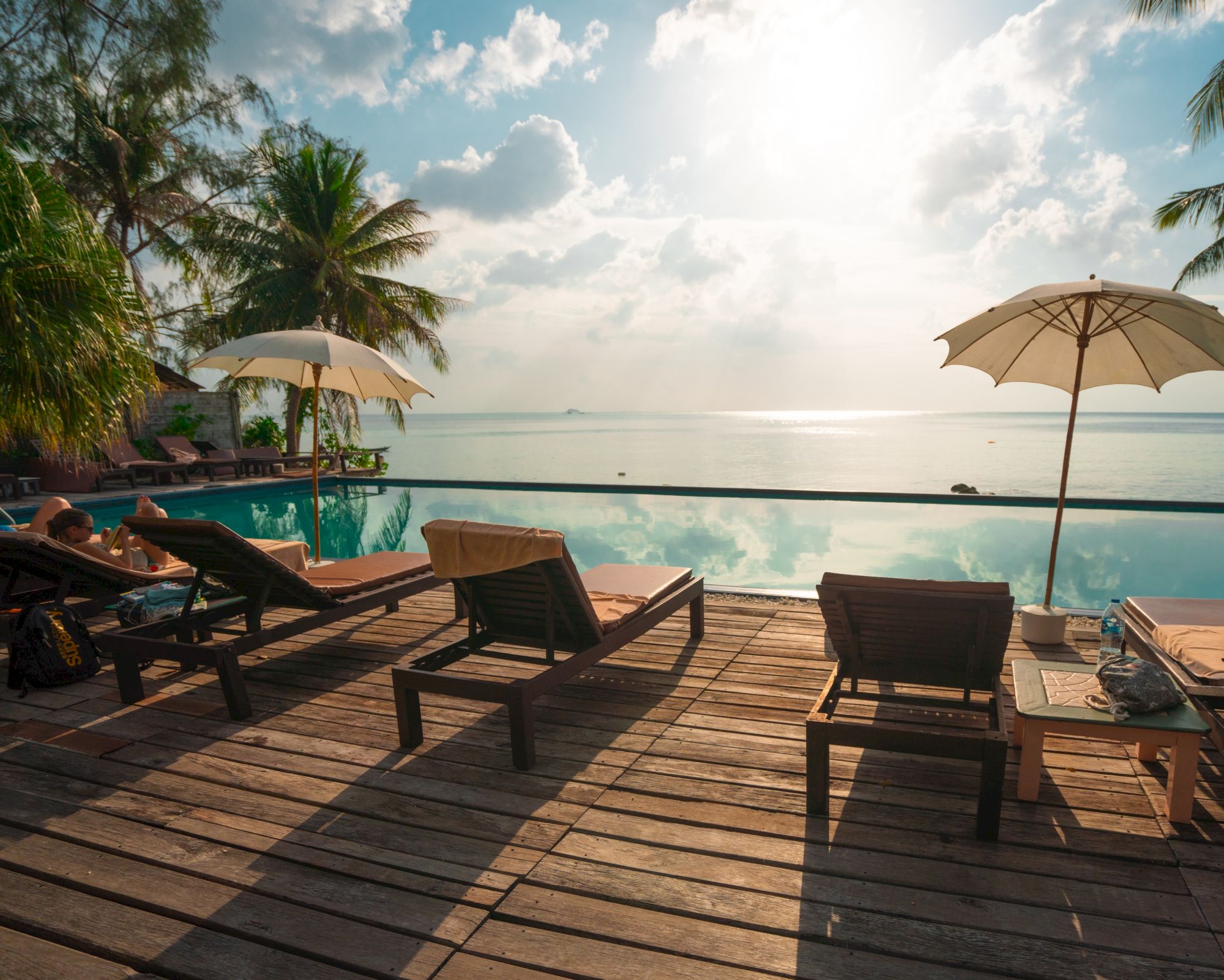 A serene poolside scene with lounge chairs, umbrellas, and palm trees overlooking a calm ocean under a cloudy sky.