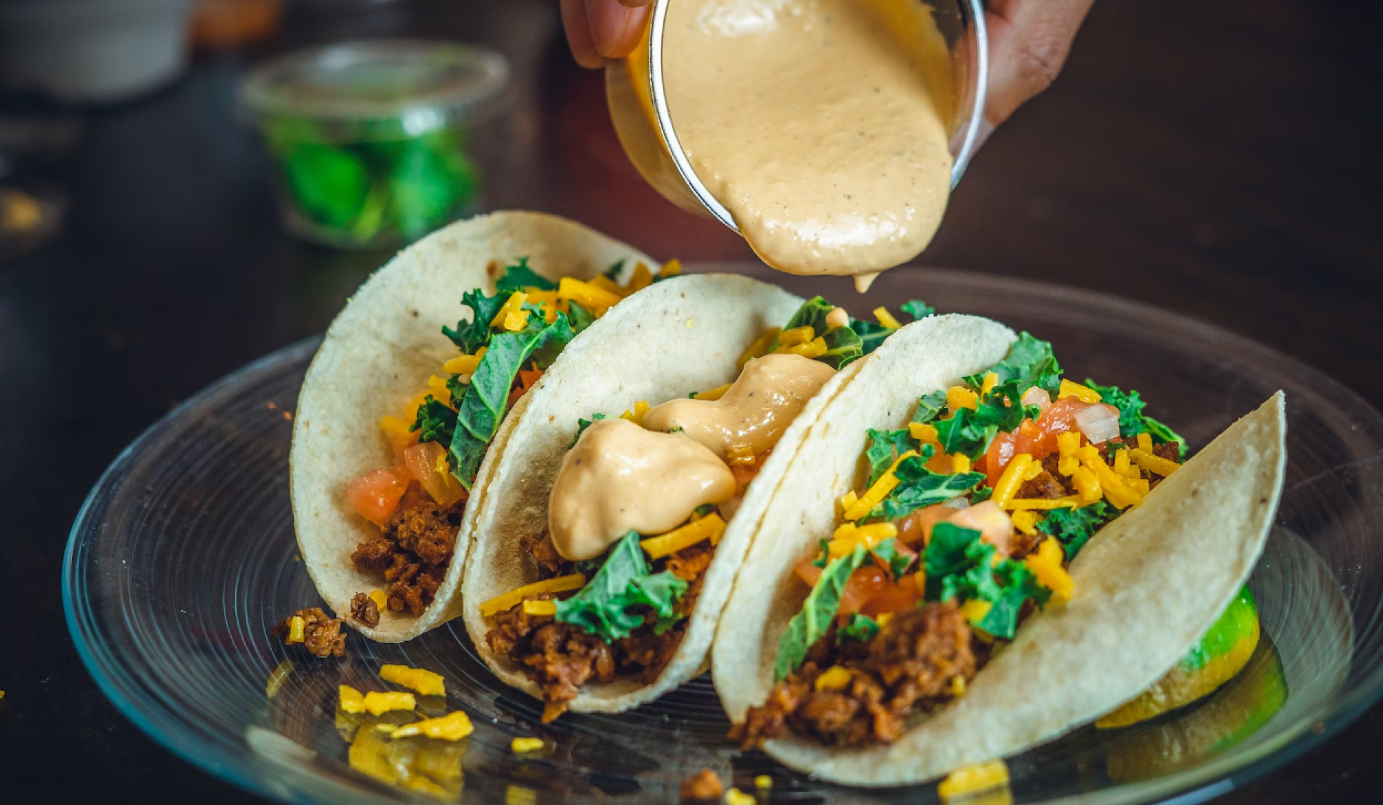 Three tacos with toppings like lettuce, cheese, and sauce being poured over them, on a glass plate.