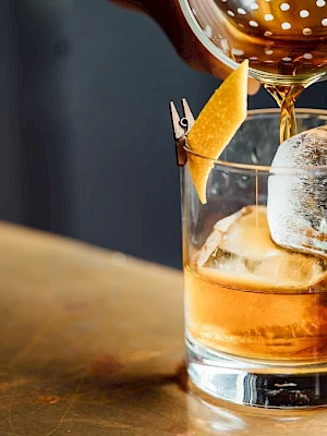 A cocktail being poured into a glass with ice and a lemon twist garnish sits on a textured countertop, captured in warm lighting.