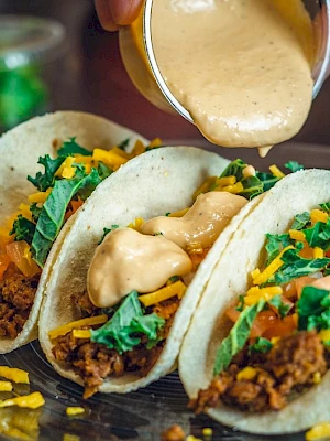 Three tacos with toppings like ground meat, cheese, and greens, being drizzled with sauce on a plate.