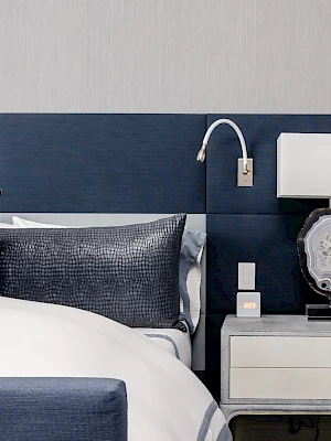 A modern bedroom features a bed with blue pillows, a nightstand, a decorative lamp, and a wall-mounted reading light.
