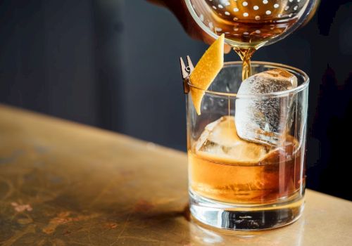 A cocktail is being poured into a glass with ice and a lemon twist garnish on a wooden surface.
