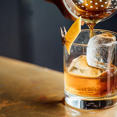 A cocktail is being poured into a glass with ice and a lemon twist garnish on a wooden surface.