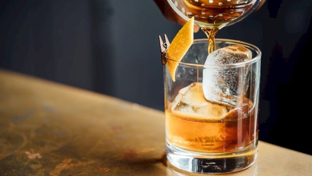 A cocktail is being poured into a glass with ice and a lemon twist garnish on a wooden surface.