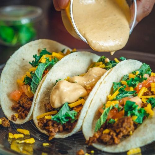 Three tacos with ground meat, lettuce, cheese, and tomatoes on a plate, being topped with a creamy sauce.