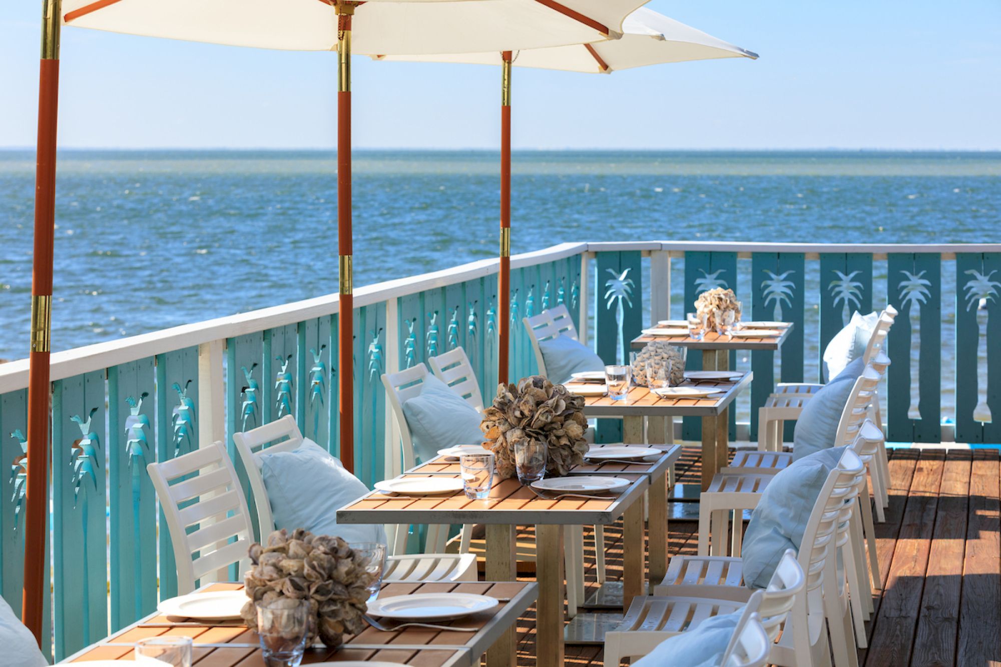 A seaside restaurant patio with tables set under umbrellas, overlooking the ocean, featuring decorative elements and a relaxed atmosphere.