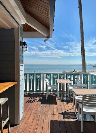 Outdoor dining area with wooden tables, chairs, and a bar counter overlooking a sunny beach and ocean, surrounded by palm trees.