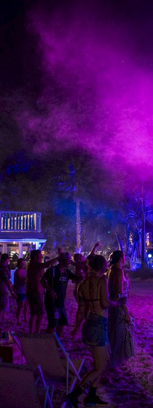 A vibrant beach party at night with people dancing under purple lights, near a two-story building and a stage.