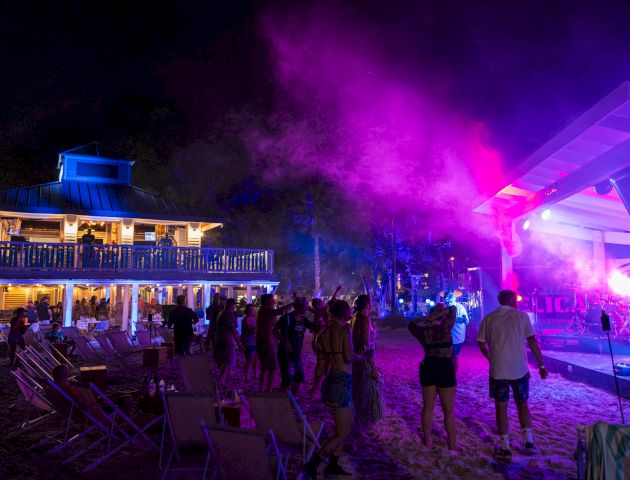 A vibrant beach party at night with people dancing under purple lights, near a two-story building and a stage.