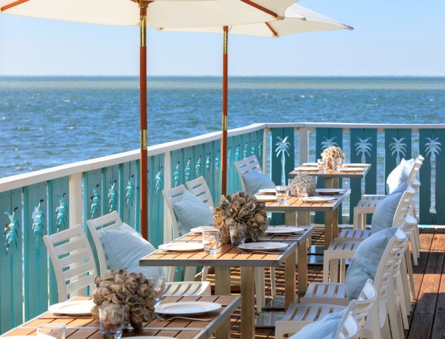 A seaside deck dining setup with white chairs, table settings, and umbrellas overlooking the ocean.