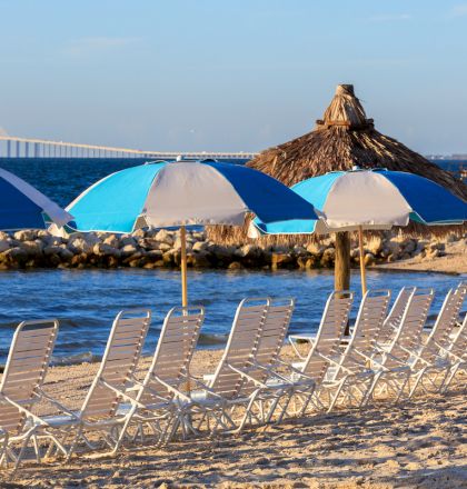 A sunny beach scene with blue umbrellas and wooden deck chairs facing the sea, calm water in the distance.