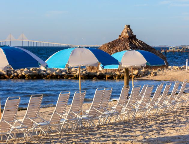 A sandy beach scene with rows of lounge chairs, blue and white umbrellas, a tiki hut, rocky shoreline, and a bridge in the background.