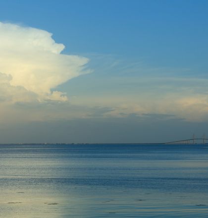 A calm seascape with a partly cloudy sky and a bridge visible on the horizon, under a blue sky.