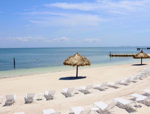 A serene beach with rows of white loungers, straw umbrellas, calm blue sea, and a wooden pier under a clear sky.