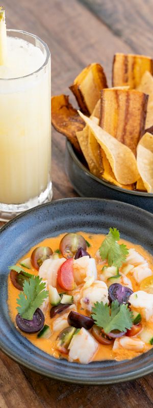 Shrimp ceviche in a bowl, lime garnish, plantain chips, and a pineapple drink cocktail on a wooden table.