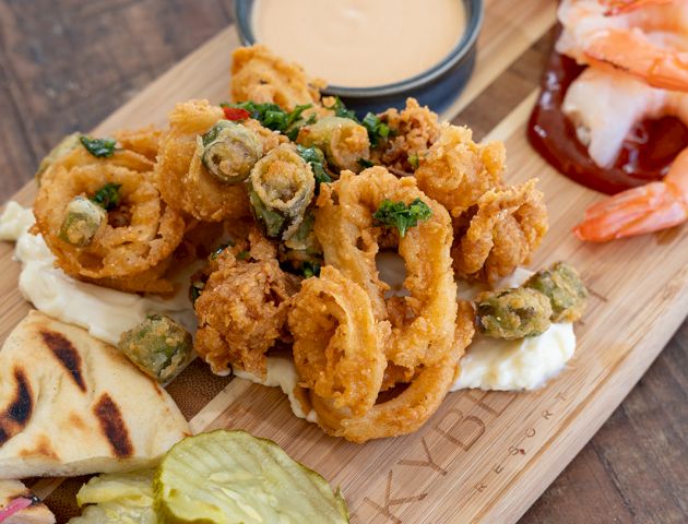 A wooden board of crispy fried rings with shrimp, a cup of creamy dipping sauce, plus pita, cucumber, pickles and salad.