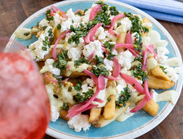 A plate of loaded fries with feta, chopped herbs, and pink pickled onions, served with cutlery on a table.