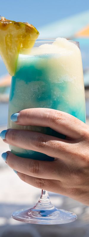 A person holds a tropical cocktail with blue and white layers topped with a pineapple slice on a beach with colorful umbrellas.