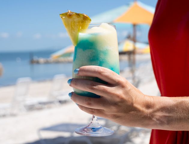 A person holds a tropical cocktail with blue and white layers topped with a pineapple slice on a beach with colorful umbrellas.