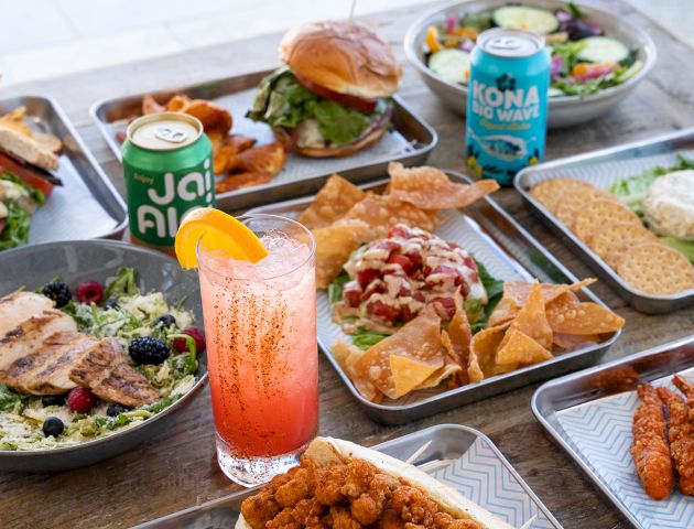An array of food and drinks, including sandwiches, salads, chicken wings, chips, and canned beverages on a table, with a pink drink.